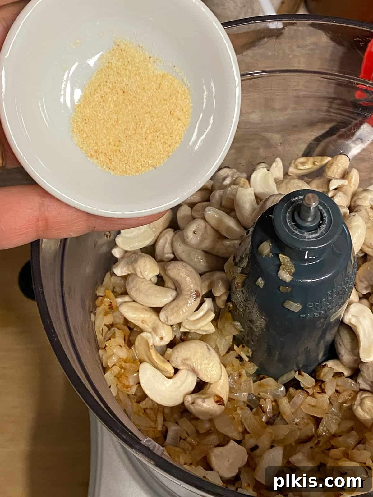 Gourmet Vegan French Onion Dip 6 Process shot: Garlic powder is being sprinkled into the food processor, joining the cashews and other ingredients.