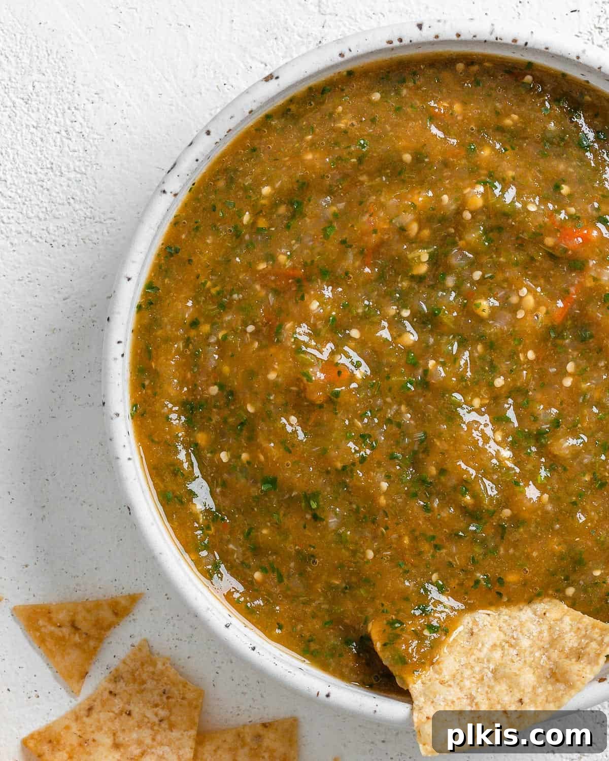 Zesty Tomatillo Green Chili Salsa 2 A bowl of vibrant green tomatillo salsa with a tortilla chip being dipped in, surrounded by scattered chips, showcasing its fresh appeal.