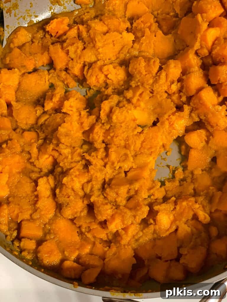 Golden Sweet Potato Rolls 9 Process shot demonstrating softened sweet potatoes being mashed with a potato masher in a pan, creating a creamy texture.