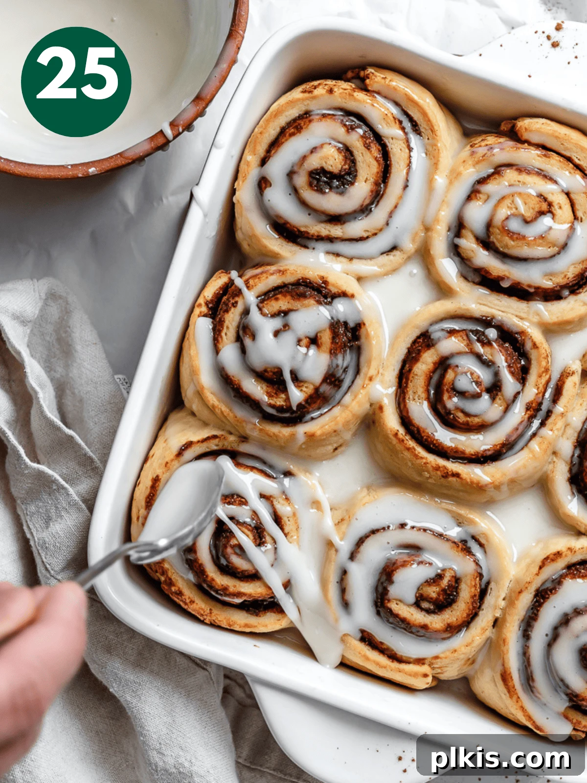 Heavenly Vegan Cinnamon Rolls 28 Vanilla icing being drizzled generously over warm, baked vegan cinnamon rolls in a white pan.