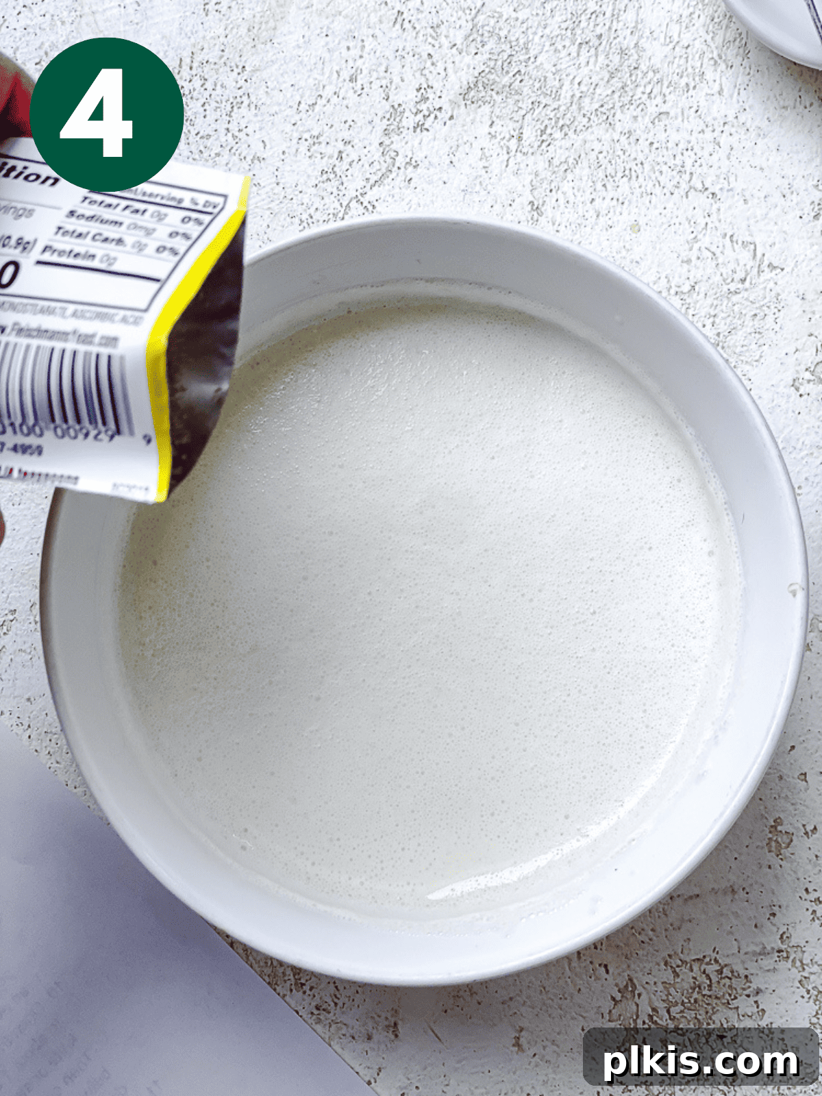 Heavenly Vegan Cinnamon Rolls 7 Pouring a packet of rapid yeast into the warm vegan buttermilk and sugar mixture in a white bowl.