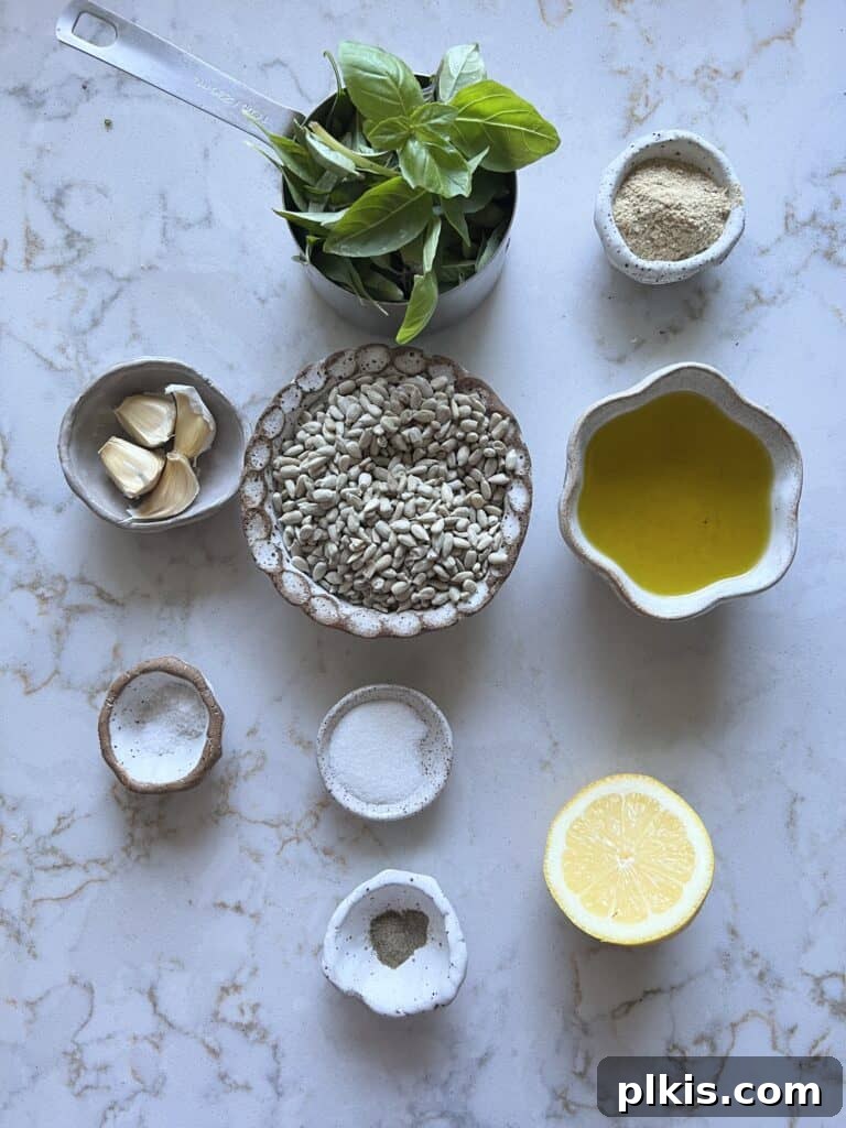 Nut-Free Vegan Basil Pesto with Sunflower Seeds 3 Ingredients for sunflower seed pesto, including fresh basil, raw sunflower seeds, garlic, olive oil, lemon, nutritional yeast, salt, pepper, and sugar, laid out on a white marble surface.