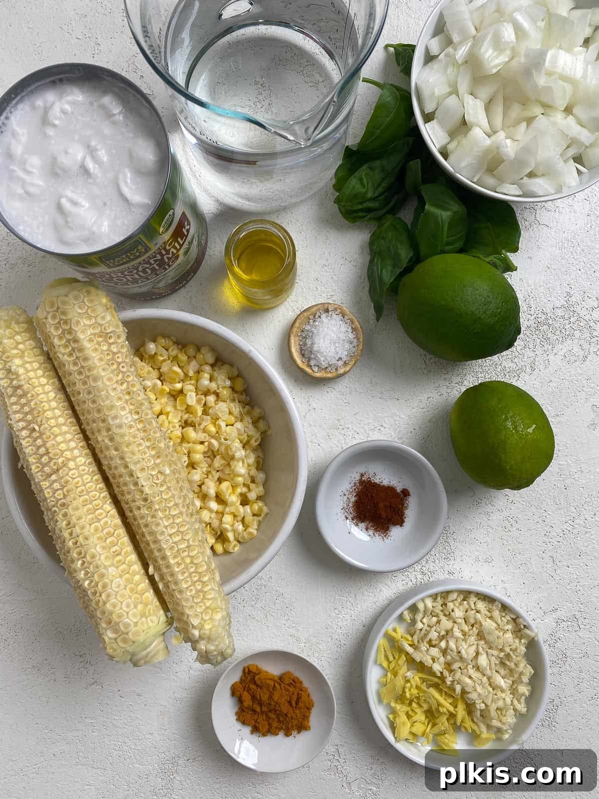 Velvety Vegan Coconut Corn Chowder 3 Assortment of fresh ingredients including corn, ginger, garlic, onion, basil, and a can of coconut milk, laid out on a white surface for vegan corn chowder.