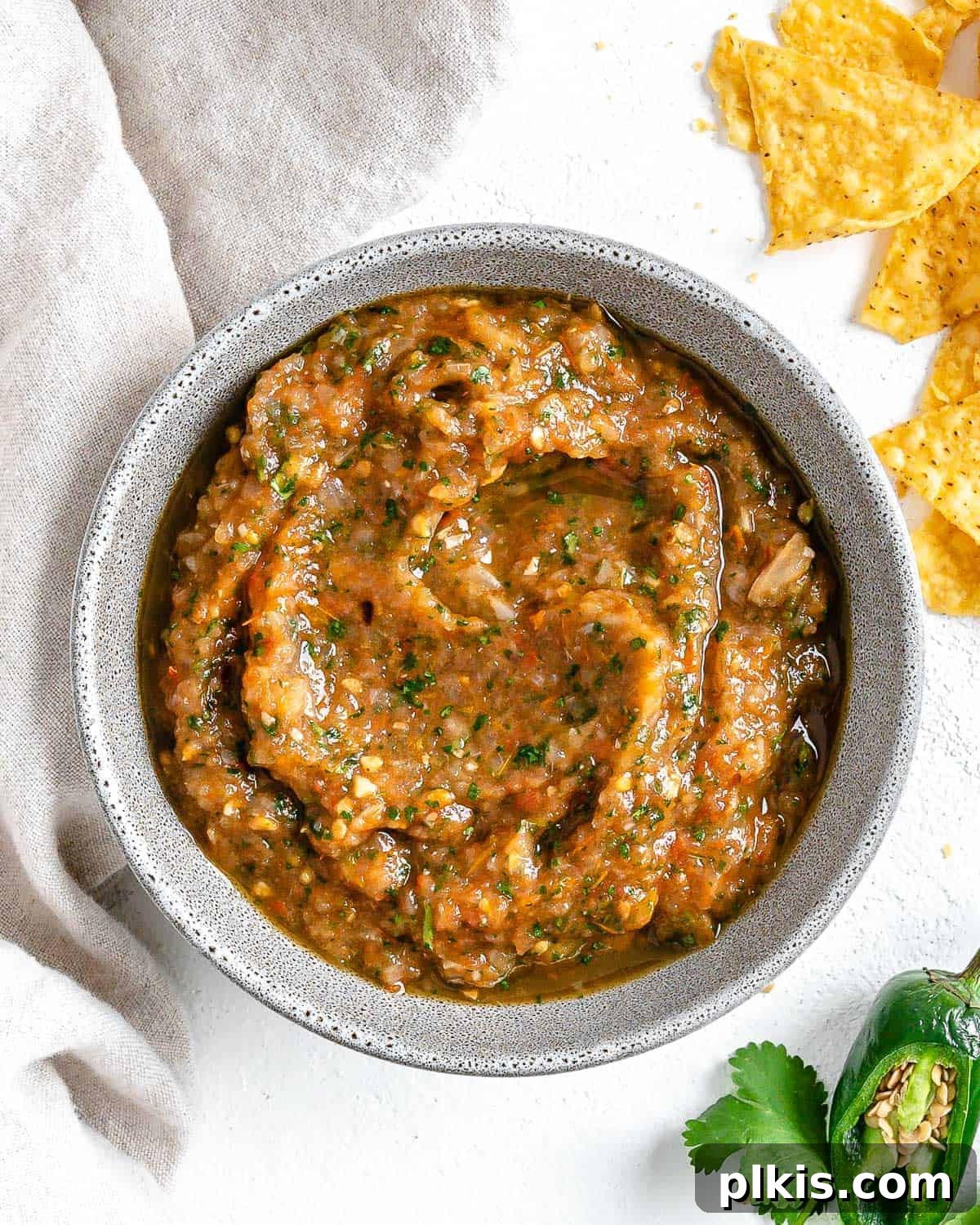 Handcrafted Roasted Red Salsa 2 A beautiful white bowl filled with Homemade Roasted Red Salsa, garnished with fresh cilantro, surrounded by crispy tortilla chips and vibrant green jalapeños on a clean white background, inviting you to dip in.