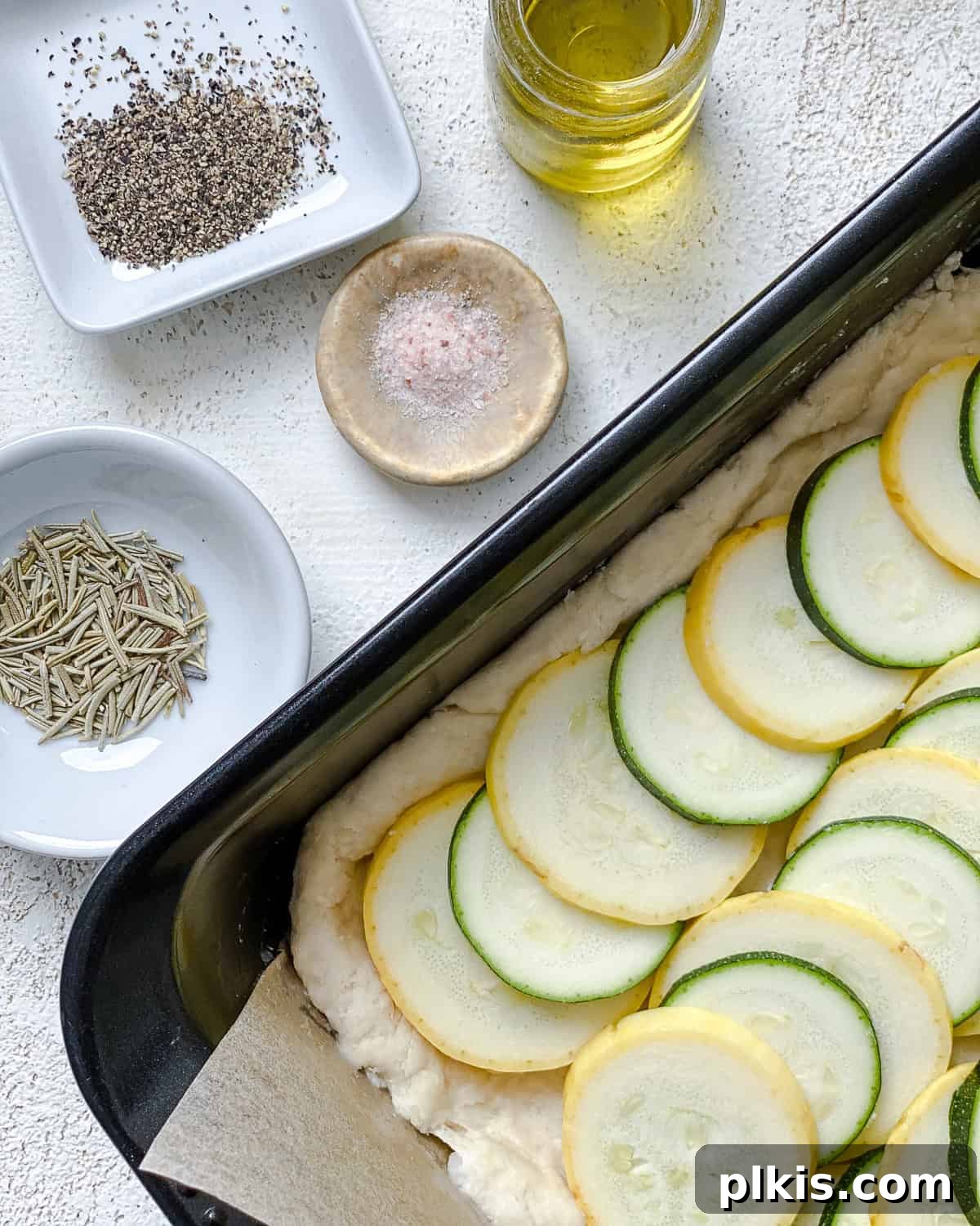 Vibrant Zucchini Tart 10 An unbaked zucchini tart in a baking dish, perfectly topped with sliced vegetables and seasonings like rosemary, olive oil, salt, and pepper.