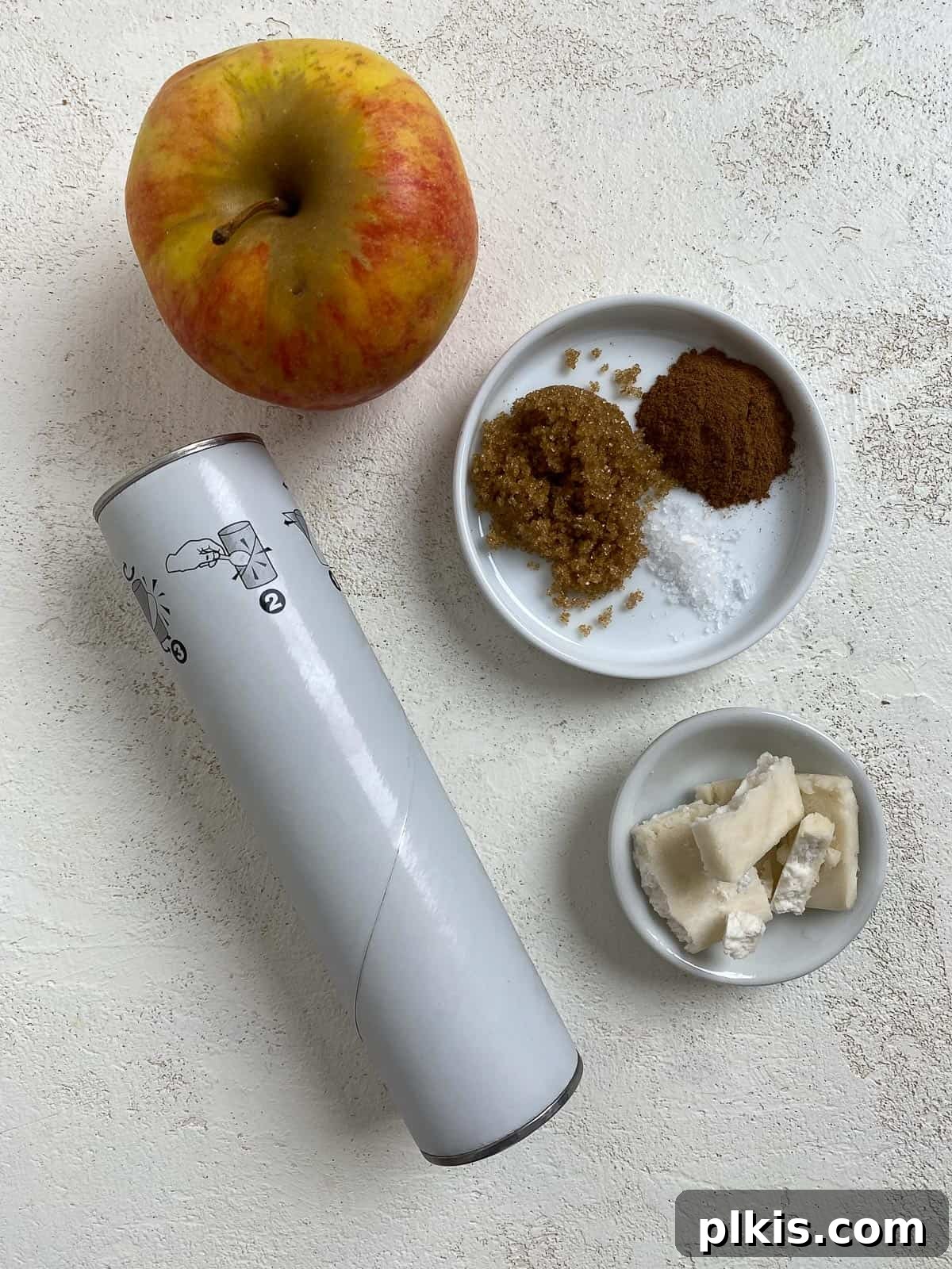 Golden Apple Cinnamon Crescents 3 Ingredients for Vegan Apple Crescent Rolls laid out on a white surface, including crescent rolls, apples, sugar, cinnamon, and vegan butter.