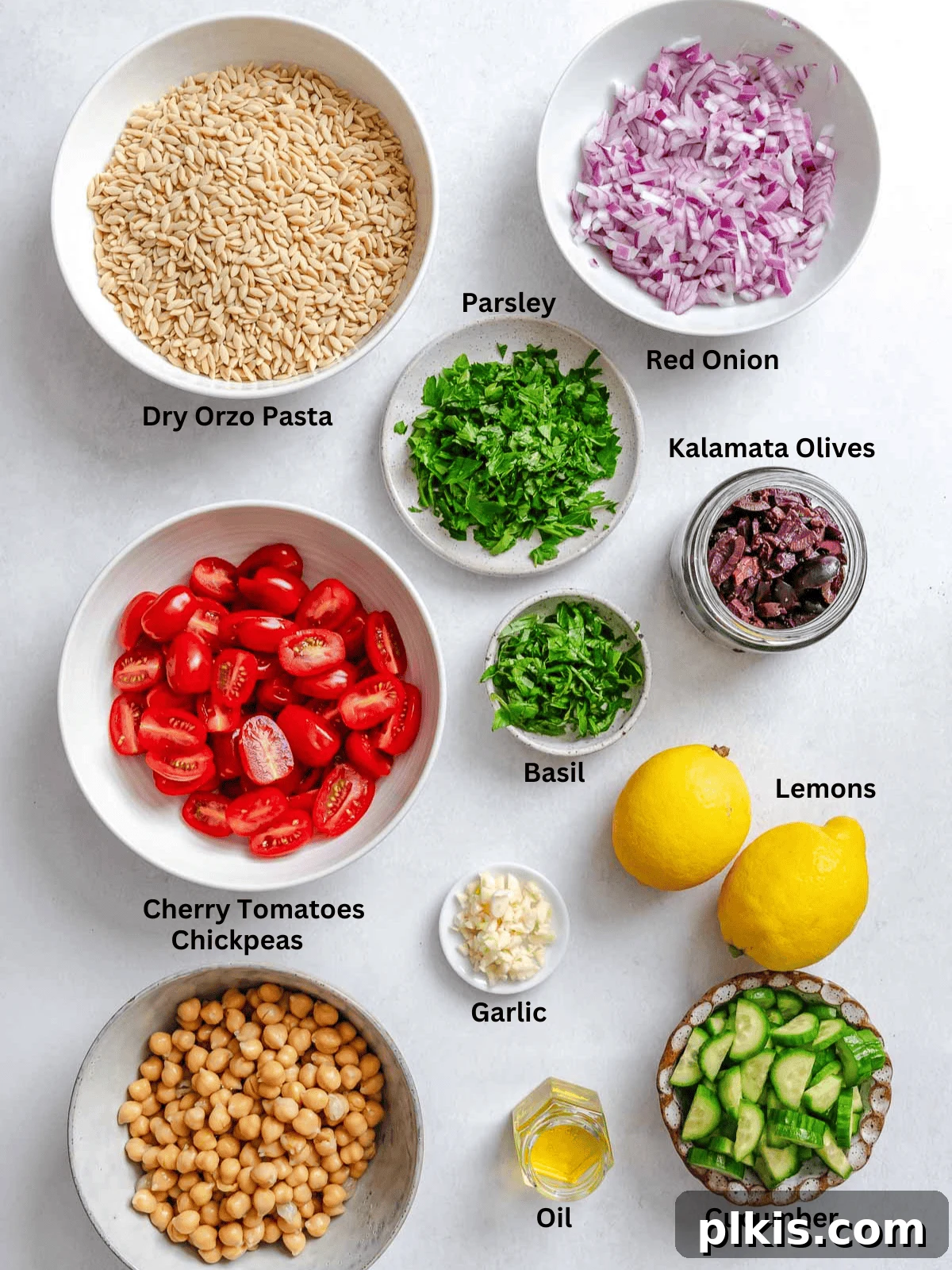 Zesty Lemon Orzo Salad 3 Various fresh ingredients for Easy Lemon Orzo Salad, including dry orzo, fresh lemons, garlic cloves, cherry tomatoes, cucumbers, red onion, chickpeas, Kalamata olives, parsley, and basil, neatly arranged on a white surface.