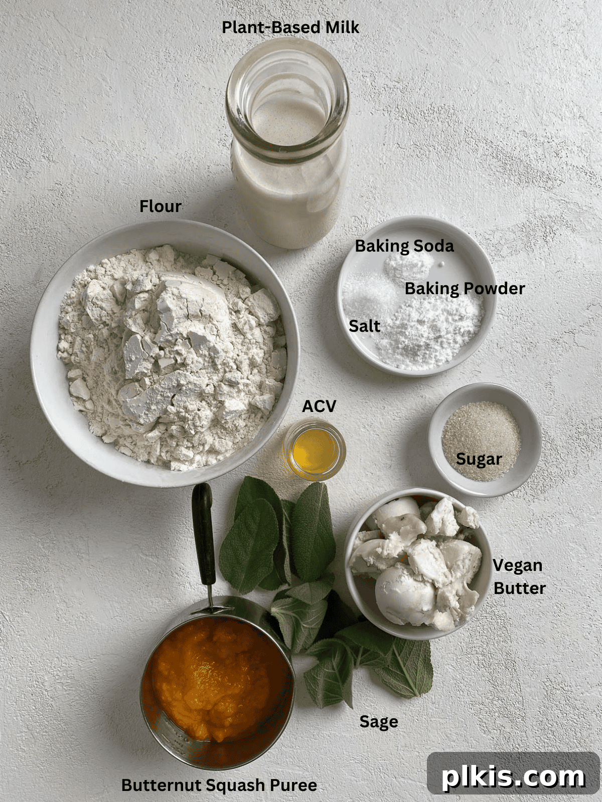 Golden Sage Butternut Biscuits 3 A selection of fresh ingredients including flour, baking powder, baking soda, sugar, salt, vegan butter, butternut squash puree, plant-based milk, apple cider vinegar, and fresh sage leaves, neatly laid out on a white surface.