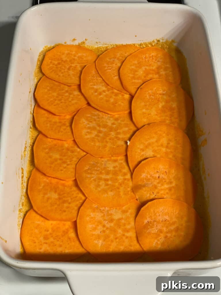Process shot: Thinly sliced sweet potatoes being carefully arranged in a single layer over the cashew cream sauce in a baking dish.