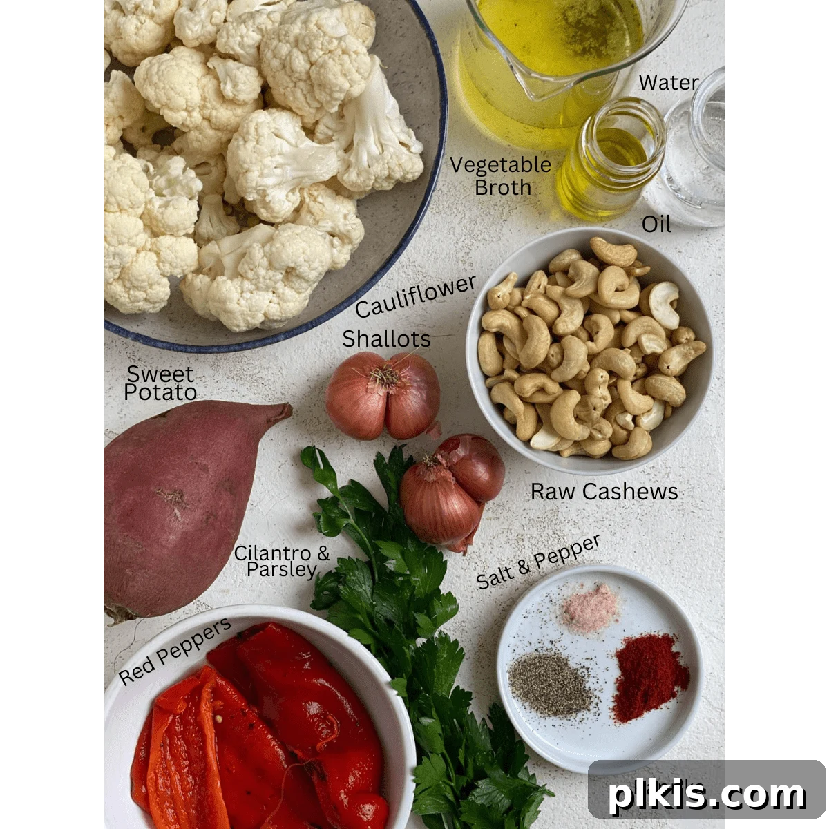 A flat lay of fresh ingredients for Sweet Potato and Cauliflower Casserole, including raw cashews, red peppers, sweet potatoes, cauliflower florets, shallots, and fresh herbs, arranged neatly on a white surface.