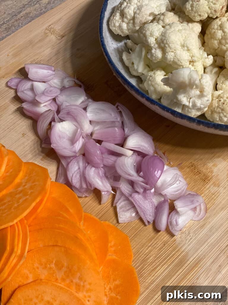 A vibrant arrangement of thinly sliced sweet potatoes, chopped shallots, and cauliflower florets on a wooden cutting board, prepped for the casserole.