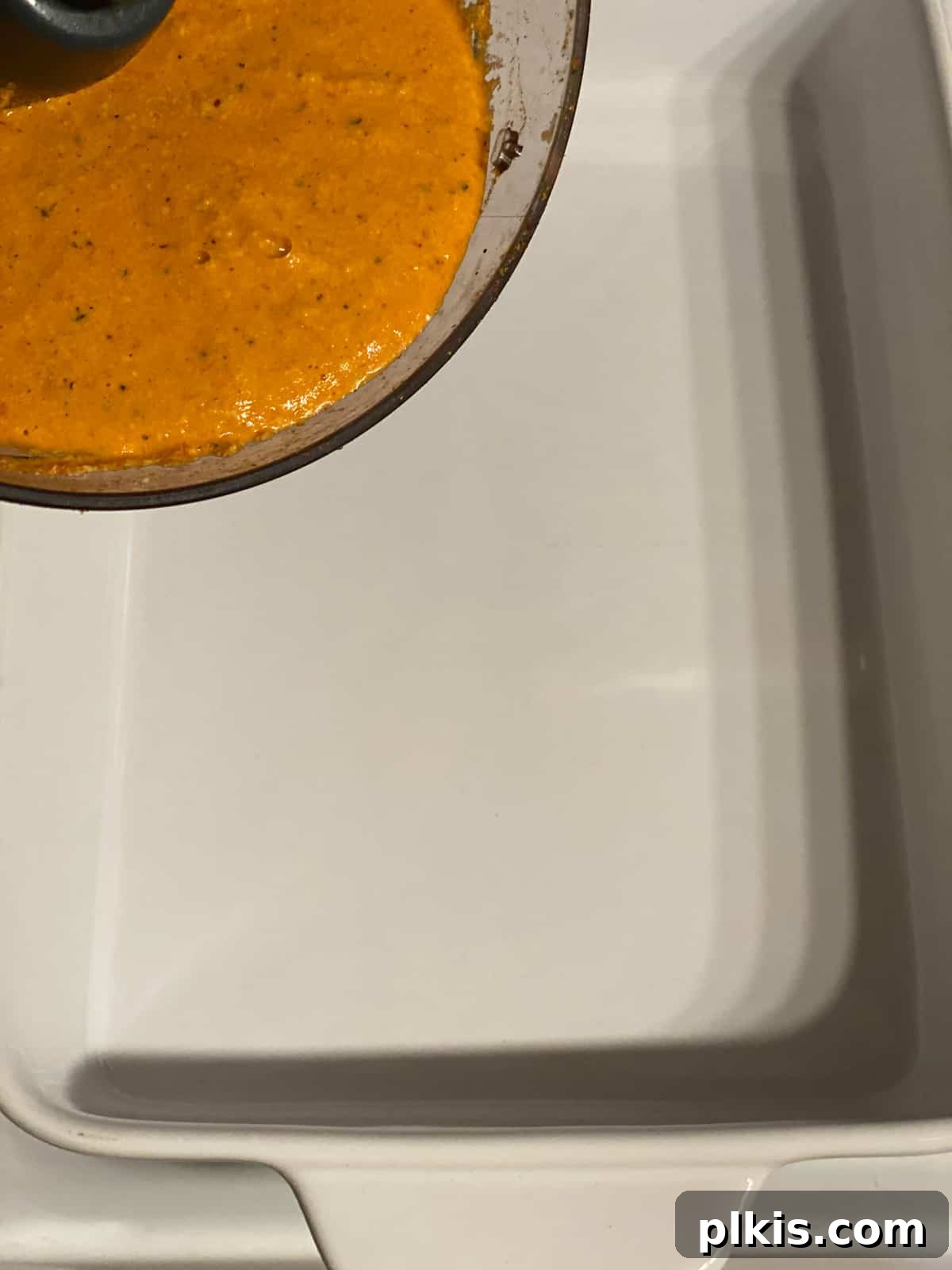 Process shot: A creamy red pepper cashew sauce being spread evenly across the bottom of a 9x13 inch baking dish, forming the first layer of the casserole.
