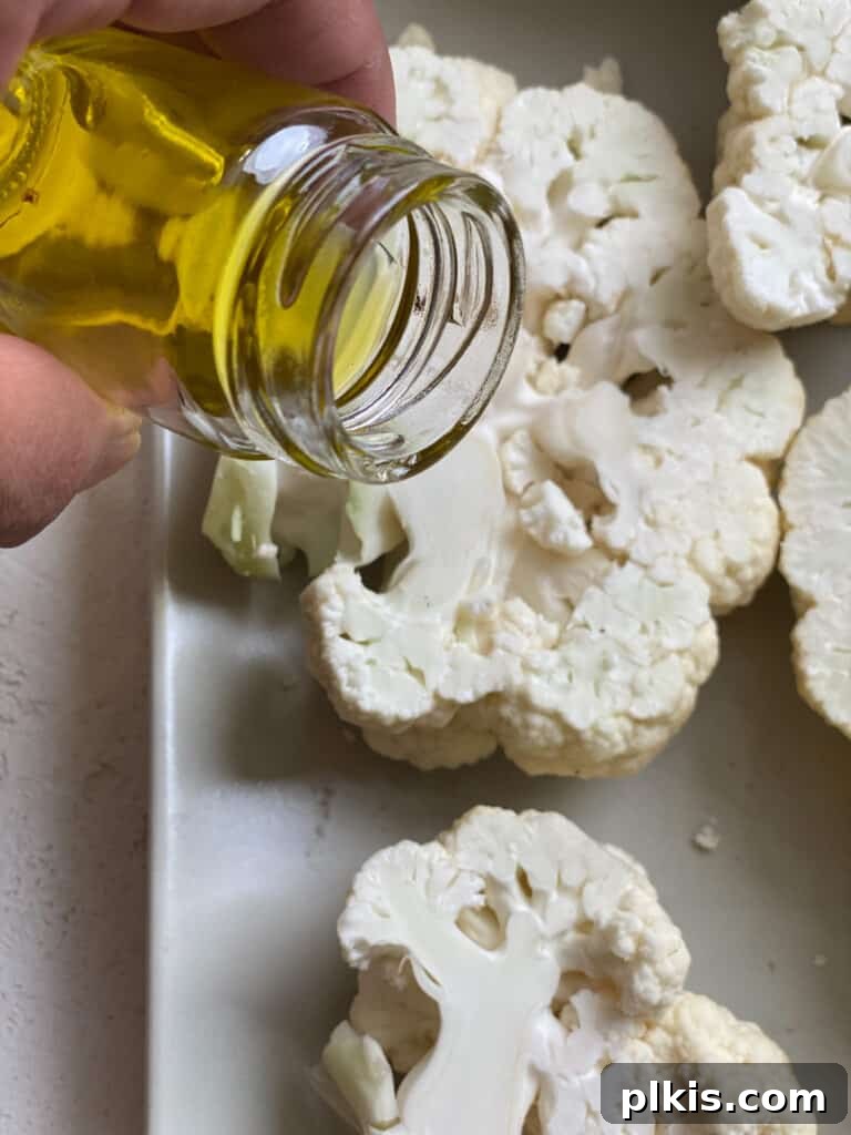 A hand gently spreading cooking oil over a cauliflower steak on a baking sheet, preparing it for roasting.