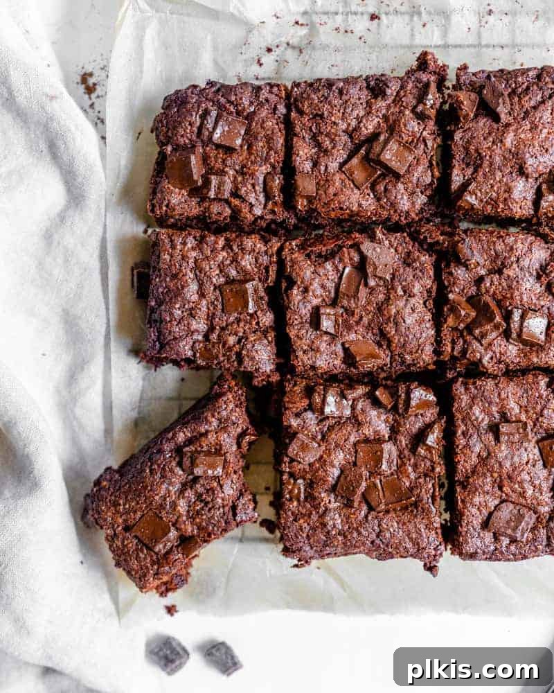 Heavenly Vegan Brownies 2 completed vegan Super Fudgy Brownies with a piece cut out on parchment paper, showcasing their rich, dark interior and crinkly top