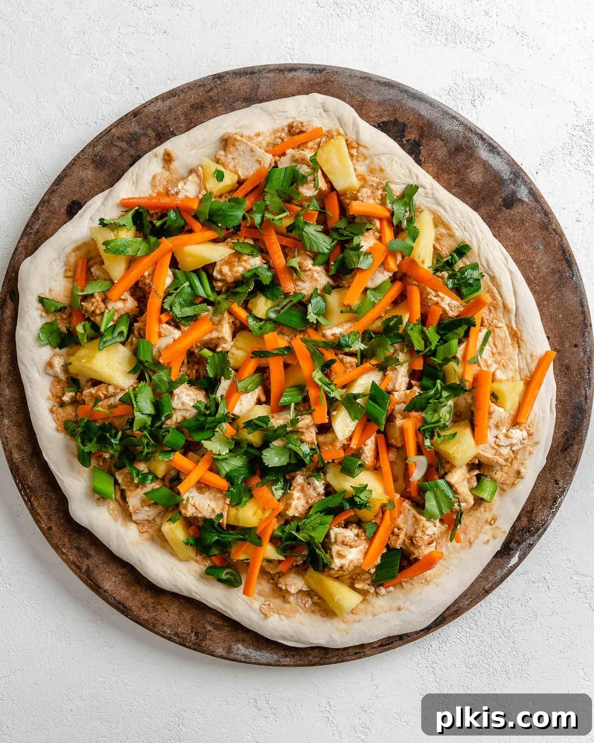 Irresistible Vegan Thai Pizza 9 process shot showing pizza toppings now placed on pizza dough against a brown and white surface