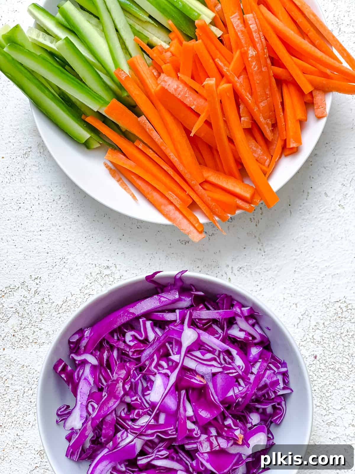 Vibrant Rainbow Rolls with Creamy Almond Shallot Dip 4 An assortment of thinly sliced vegetables for Fresh Rainbow Rolls with Almond Shallot Sauce, including cucumbers, carrots, avocado, and red cabbage, arranged on a white cutting board.