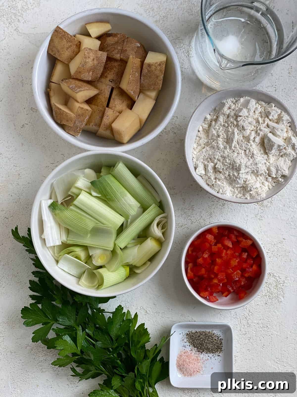 Crispy Vegan Potato Pancakes 3 A selection of fresh ingredients including chopped potatoes, leeks, flour, bell pepper, and parsley laid out on a white surface, ready for making vegan potato pancakes.
