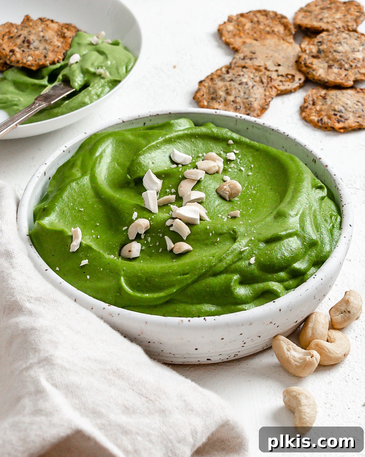 Dreamy Vegan Spinach Dip 13 A bowl of creamy, vibrant vegan spinach dip, perfectly blended and garnished with a sprinkle of fresh paprika and a few green herbs, surrounded by an assortment of crunchy crackers and fresh vegetable sticks on a rustic wooden board, ready for serving.