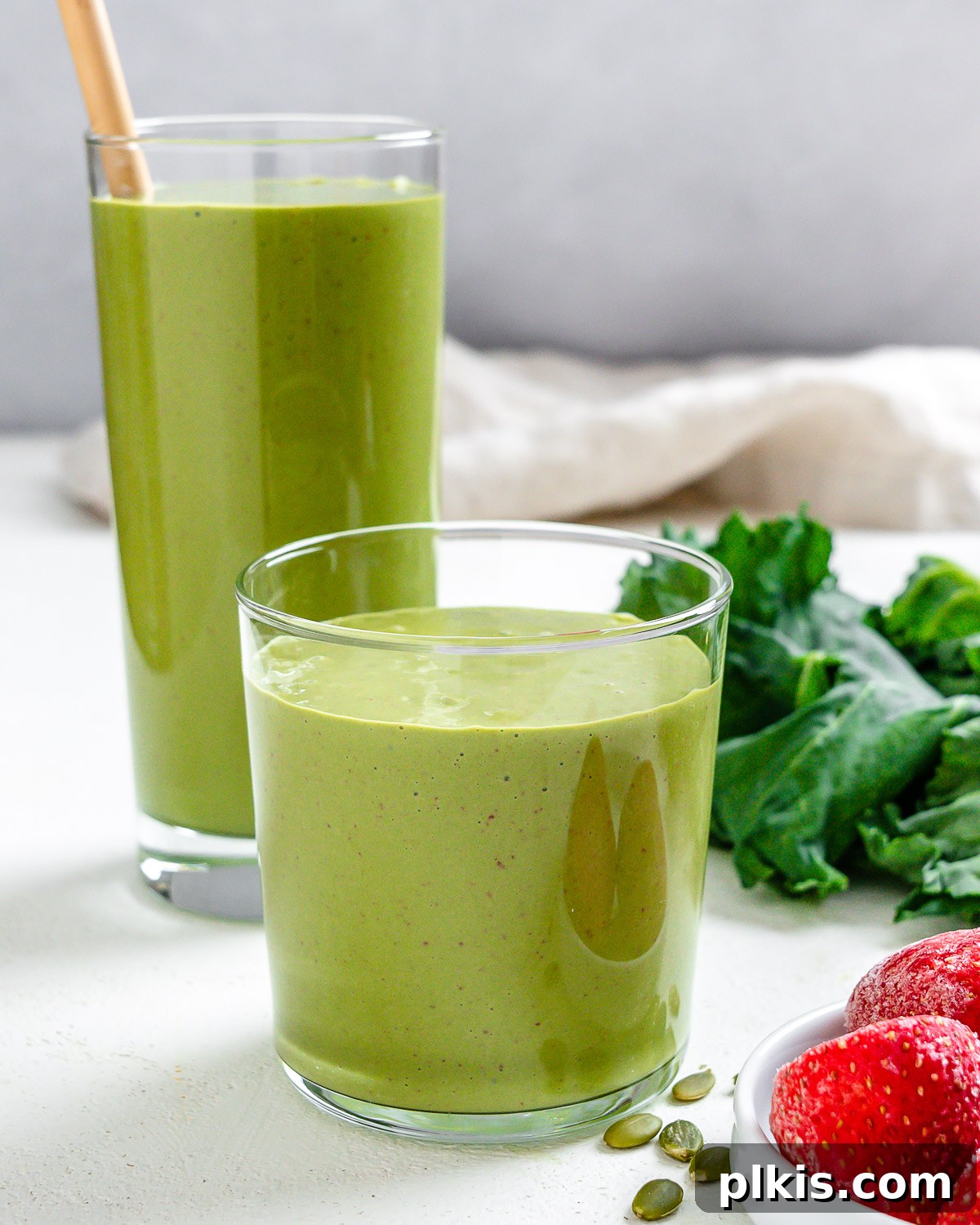 Strawberry Kale Power Elixir 2 Two glasses of vibrant Strawberry Kale Smoothie, garnished with a fresh strawberry and kale leaf, with fresh ingredients blurred in the background.