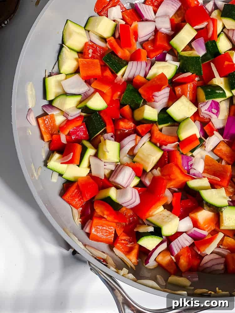 Process shot showing colorful diced vegetables, including onions, zucchini, and bell peppers, actively cooking in a large skillet on the stovetop