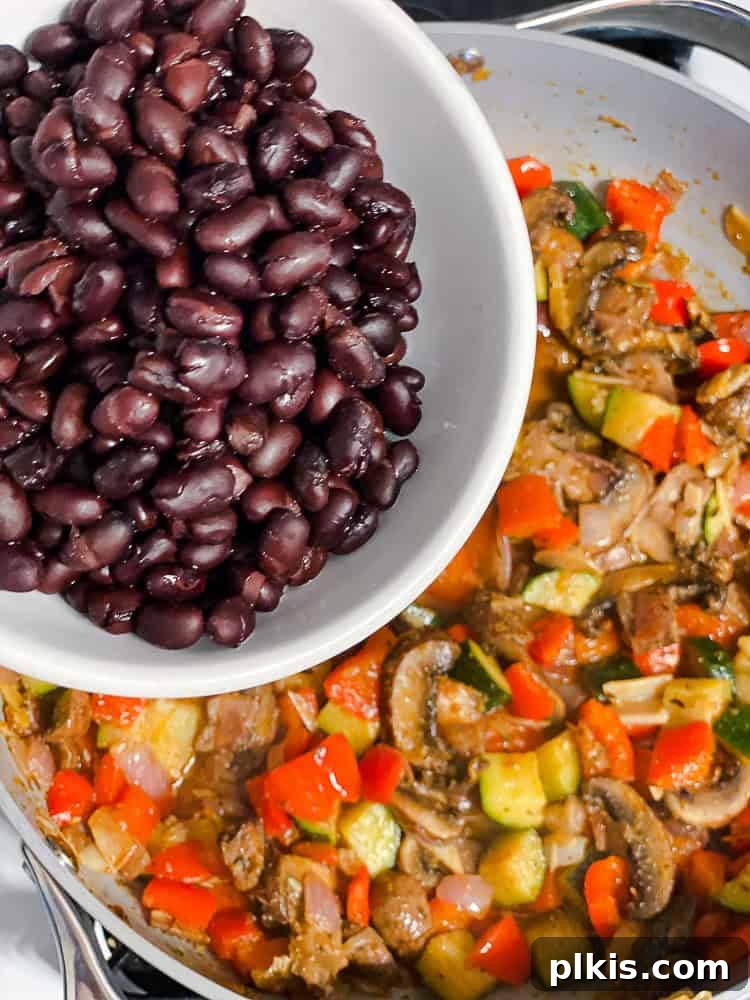 Process shot showing black beans being added to the skillet with cooked vegetables, ready to be mixed and heated through