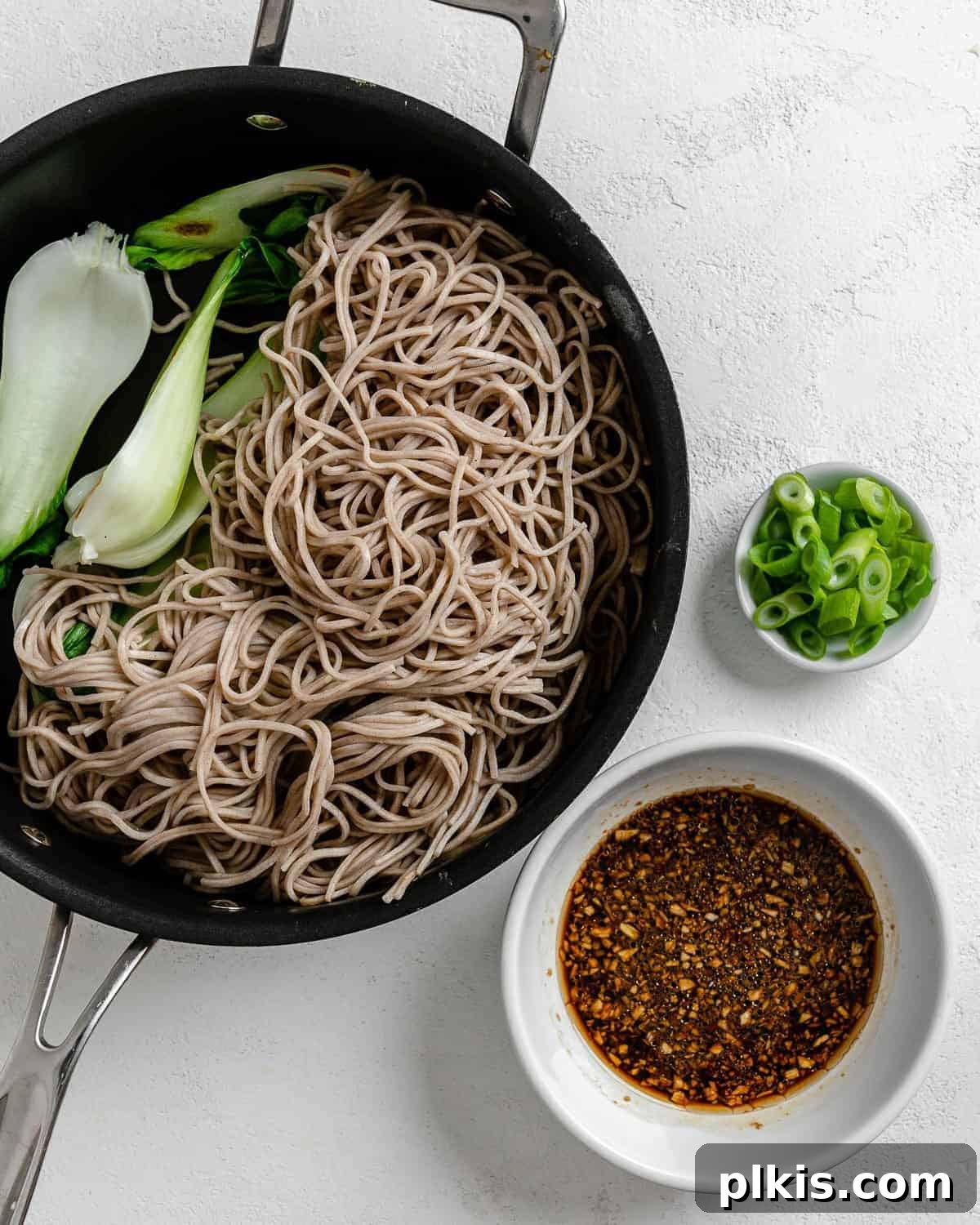 Savory Garlic Sesame Soba 6 The process of adding cooked soba noodles and vibrant green vegetables to a black pan, ready for the sauce, captured against a clean white background.