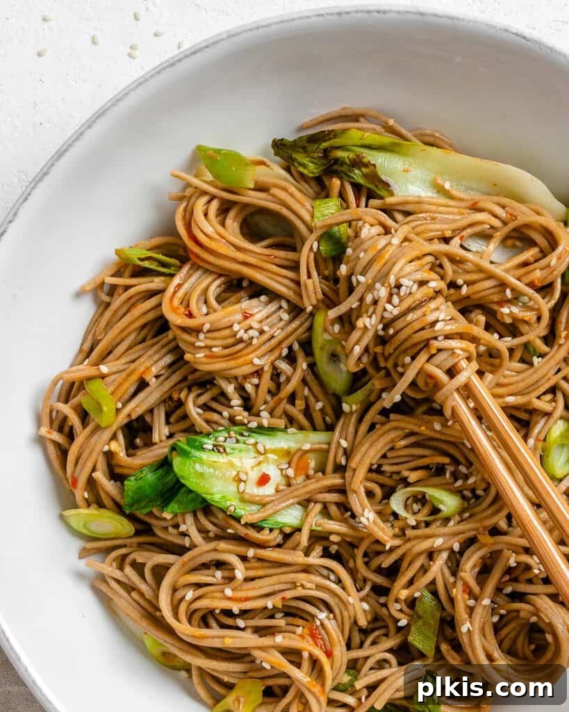 Savory Garlic Sesame Soba 8 A perfectly portioned serving of Garlic Sesame Soba Noodles in a white bowl, garnished with green onions and sesame seeds, with chopsticks resting on the rim, ready to be enjoyed.