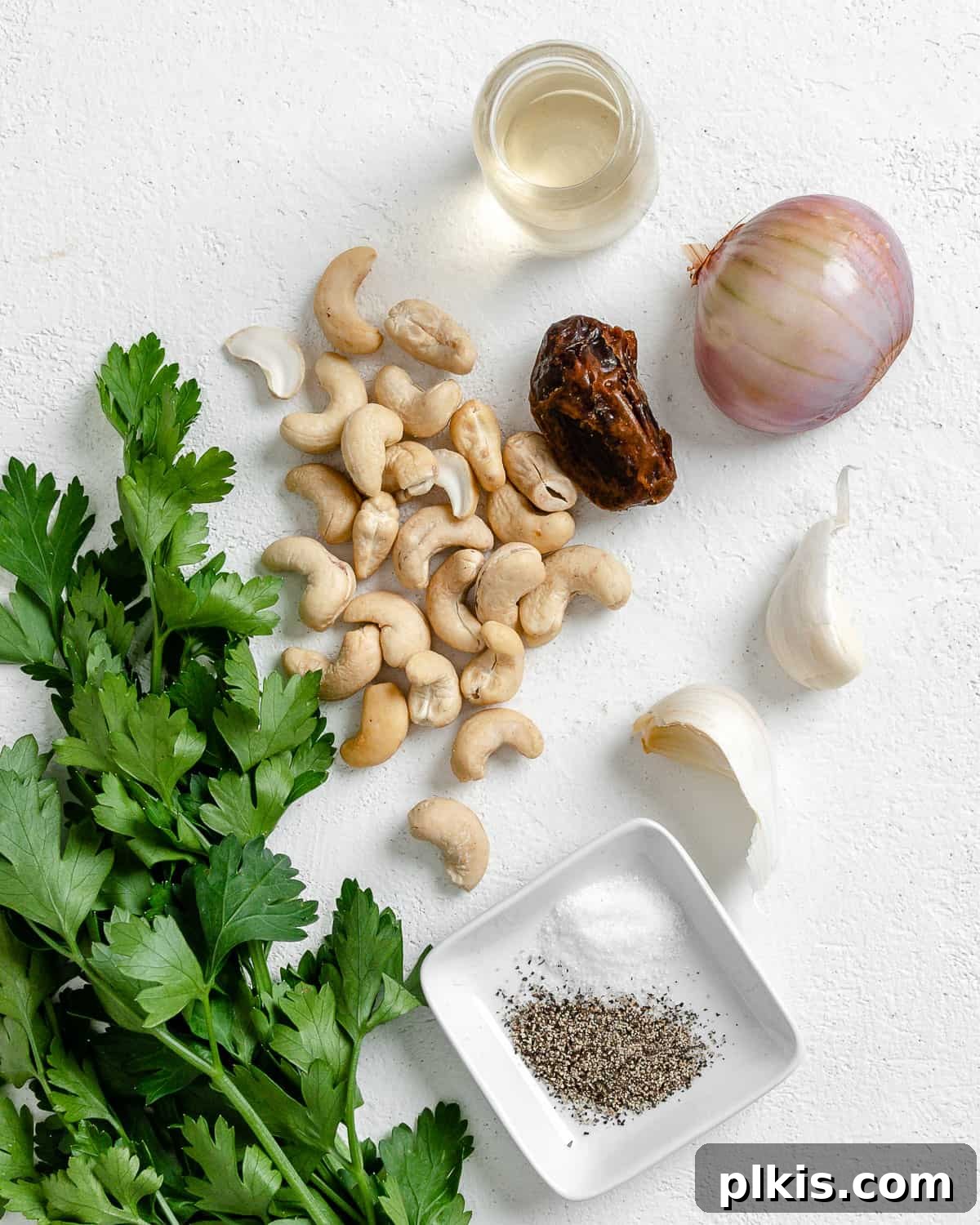 Ingredients for Creamy Vegan Garlic Sauce laid out on a white background, including fresh garlic, shallot, dates, and nuts.