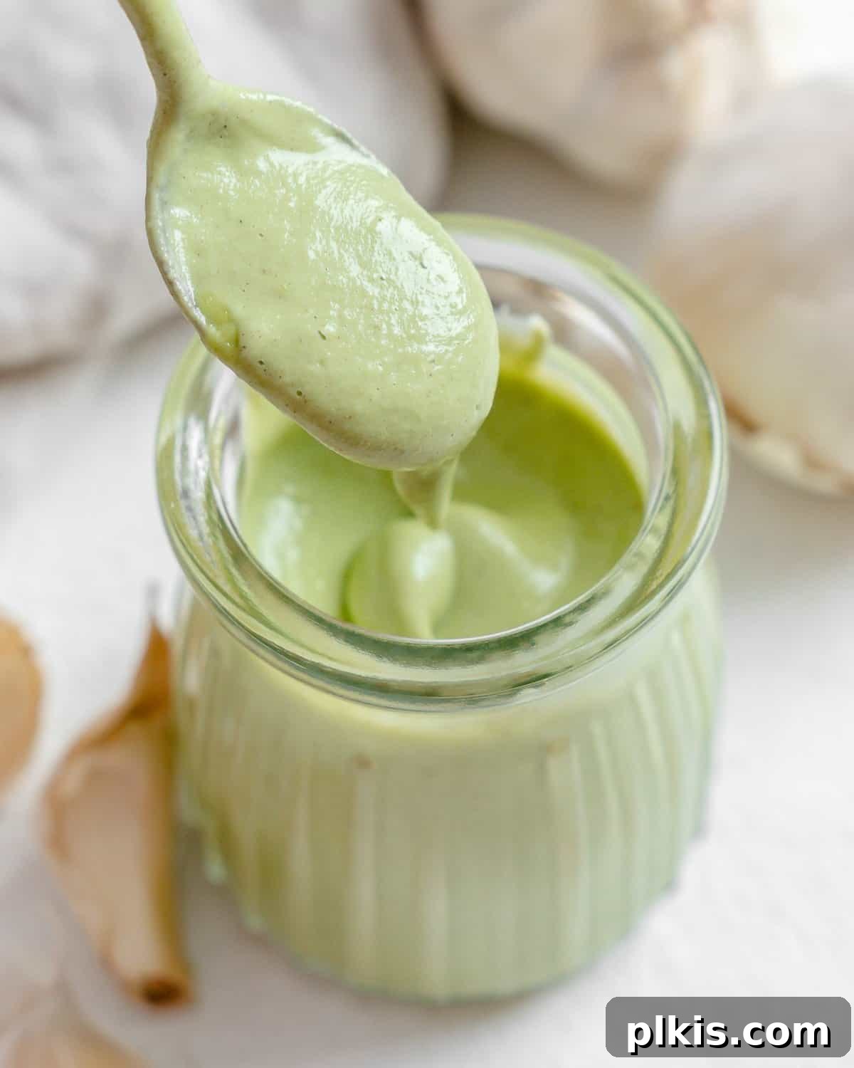 Completed Creamy Vegan Garlic Sauce in a glass jar, with a spoonful showing its thick, smooth consistency, and fresh garlic in the background.