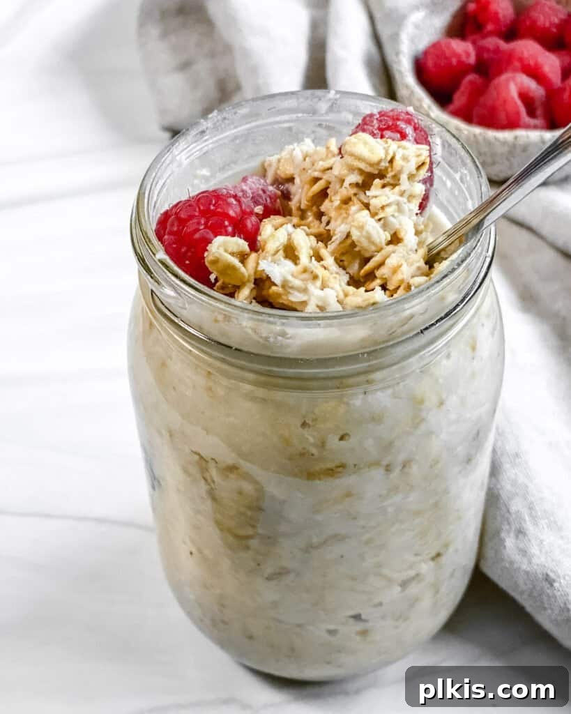 Tropical Coconut Overnight Oats 2 A beautifully arranged mason jar of Coconut Overnight Oats, generously topped with crunchy granola, an assortment of fresh berries, and banana slices, perfectly poised against a clean, white backdrop. The vibrant colors and enticing textures highlight this nutritious, plant-based breakfast.