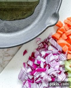 The Flavor Foundation 6 Diced mirepoix ingredients—carrots, celery, and onions—with a splash of cooking oil, resting in a black skillet before cooking.