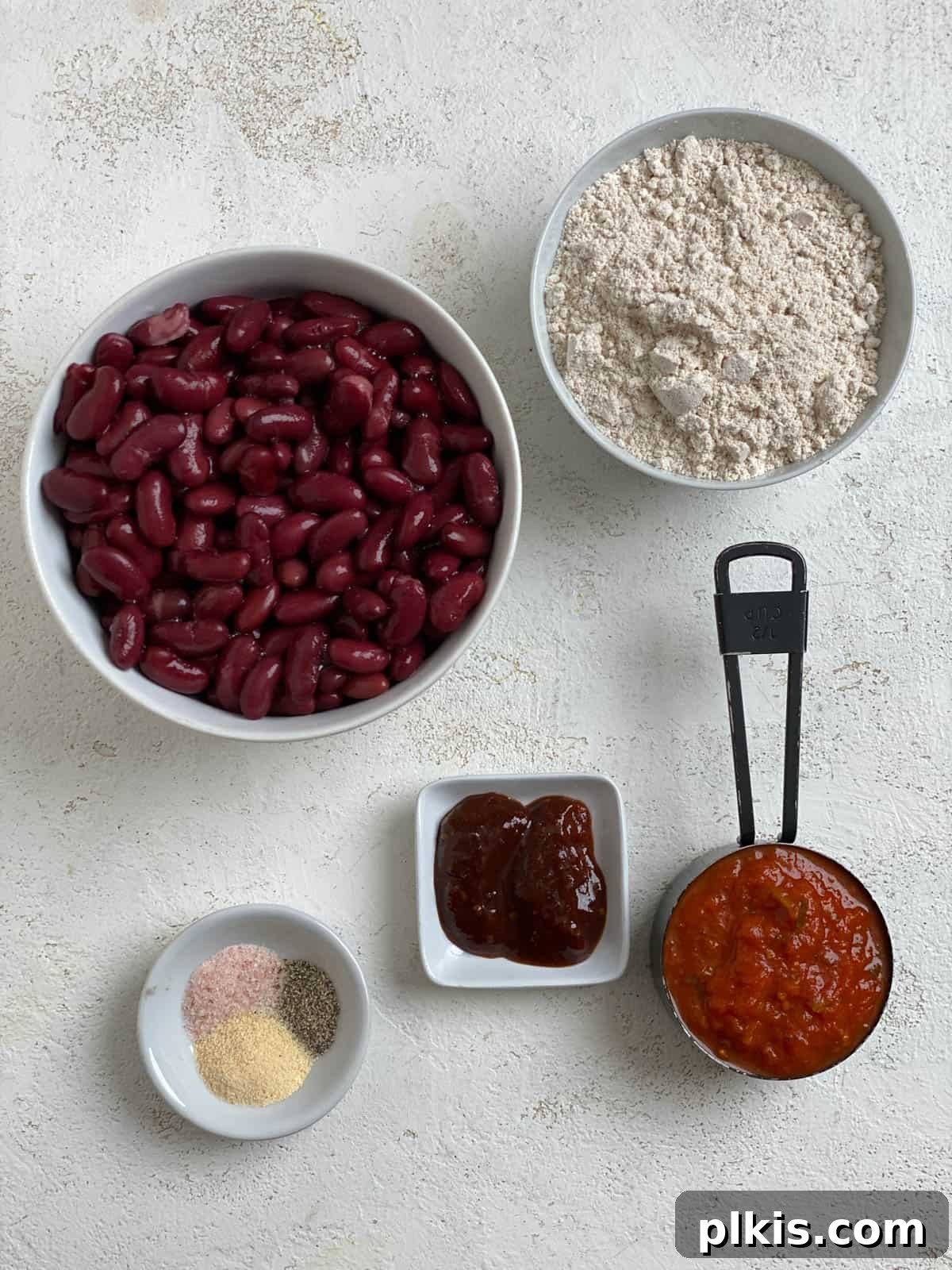 Quick-Fix 4-Ingredient Vegan Burgers 3 Four key ingredients for Easy Vegan Burger recipe measured out on a white surface: kidney beans, oat flour, sriracha, and pasta sauce.