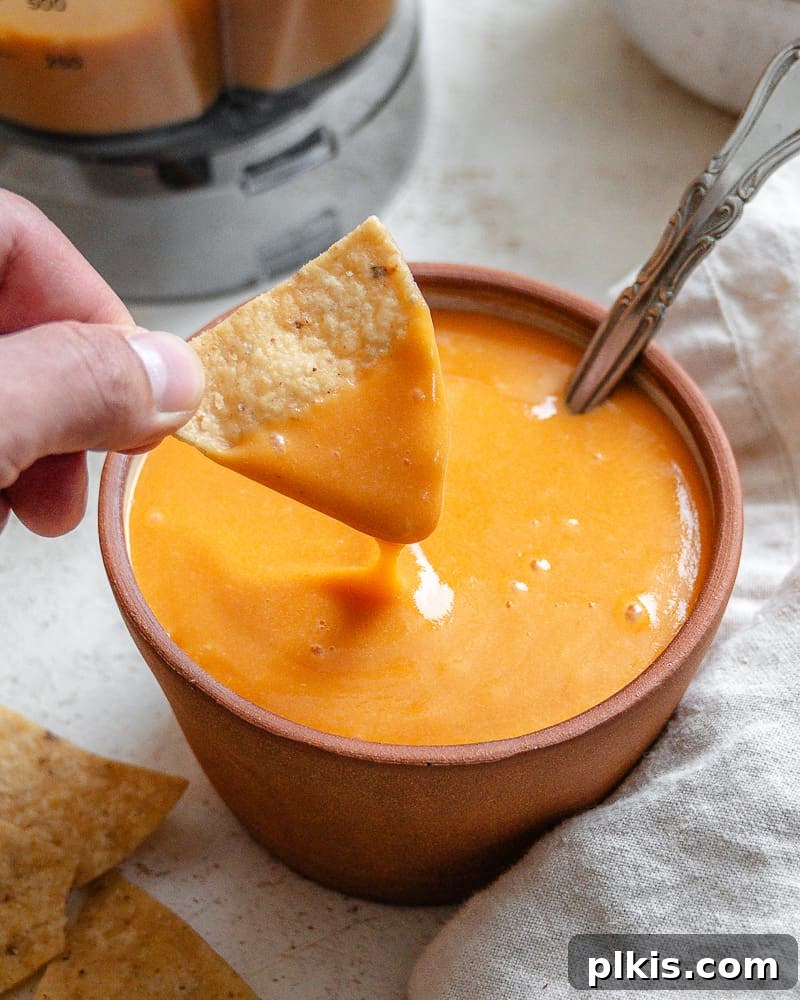 Rich Nacho Cheese Sauce 2 Hand dipping a crispy tortilla chip into a small, vibrant bowl of homemade vegan nacho cheese sauce, showcasing its rich, creamy texture.
