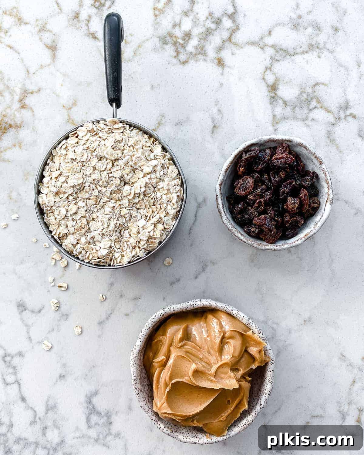 A crisp flat-lay photograph showcasing the three core ingredients: a bowl of rolled oats, a jar of creamy peanut butter, and a small pile of raisins, neatly measured out on a white surface, ready for mixing.