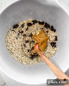 A hand mixing the oats, peanut butter, and raisins together in a white bowl, showing the sticky texture starting to form.