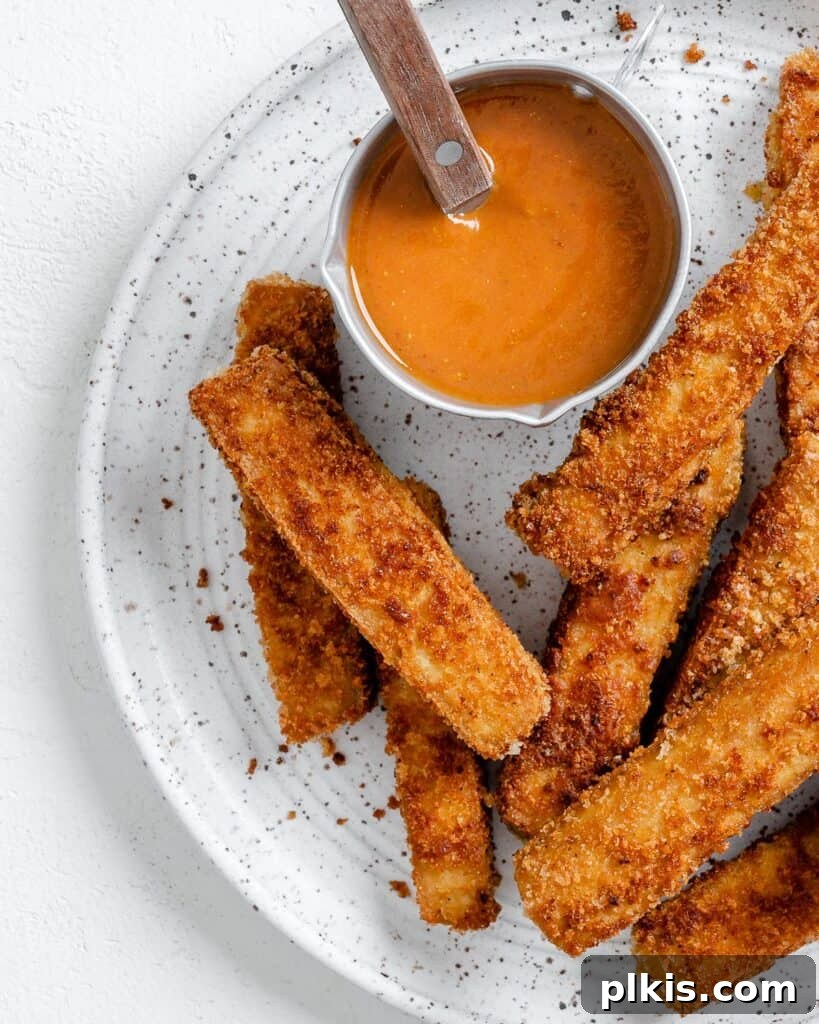 Golden-brown, crispy vegan breaded tofu sticks served on a white plate with a side of dipping sauce.