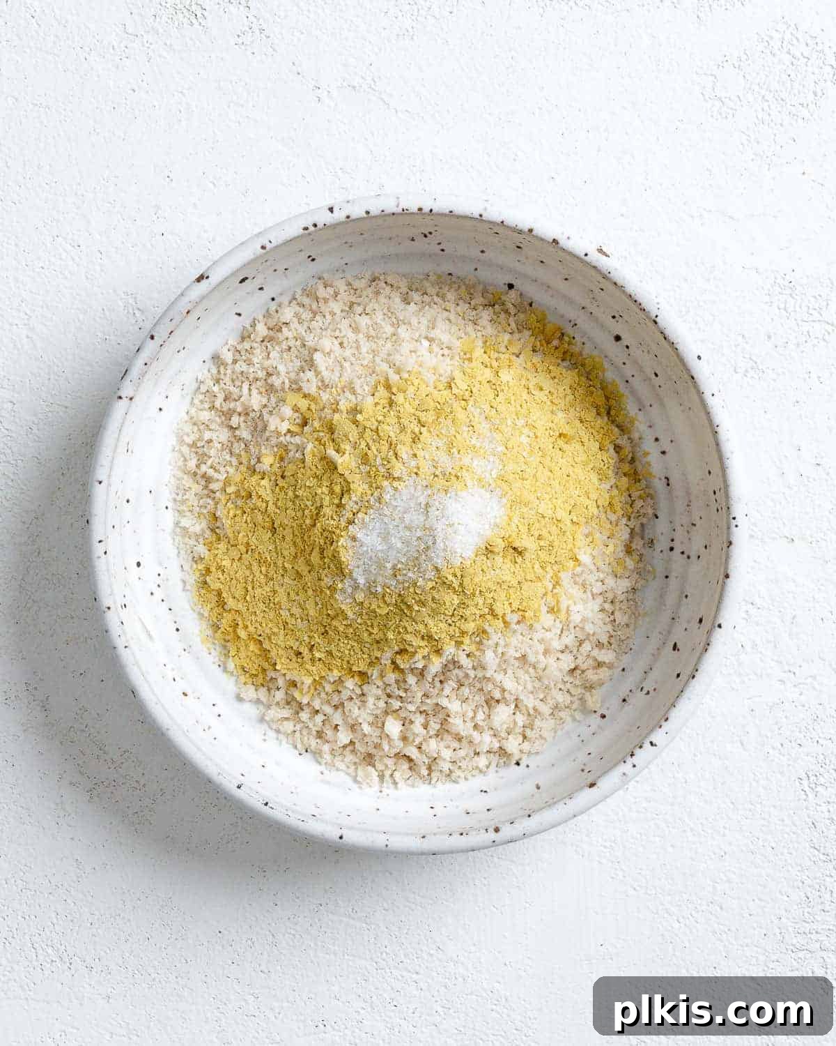 A fork mixing breadcrumbs, nutritional yeast, and salt on a white plate to create the breading mixture.