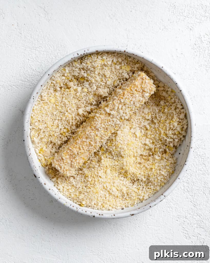 A tofu strip being pressed into a plate of breadcrumbs to achieve a complete coating.