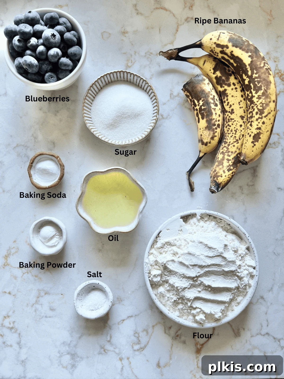 Effortless Vegan Blueberry Banana Delights 3 All ingredients for vegan blueberry banana muffins, including flour, sugar, oil, bananas, and blueberries, laid out on a white surface.