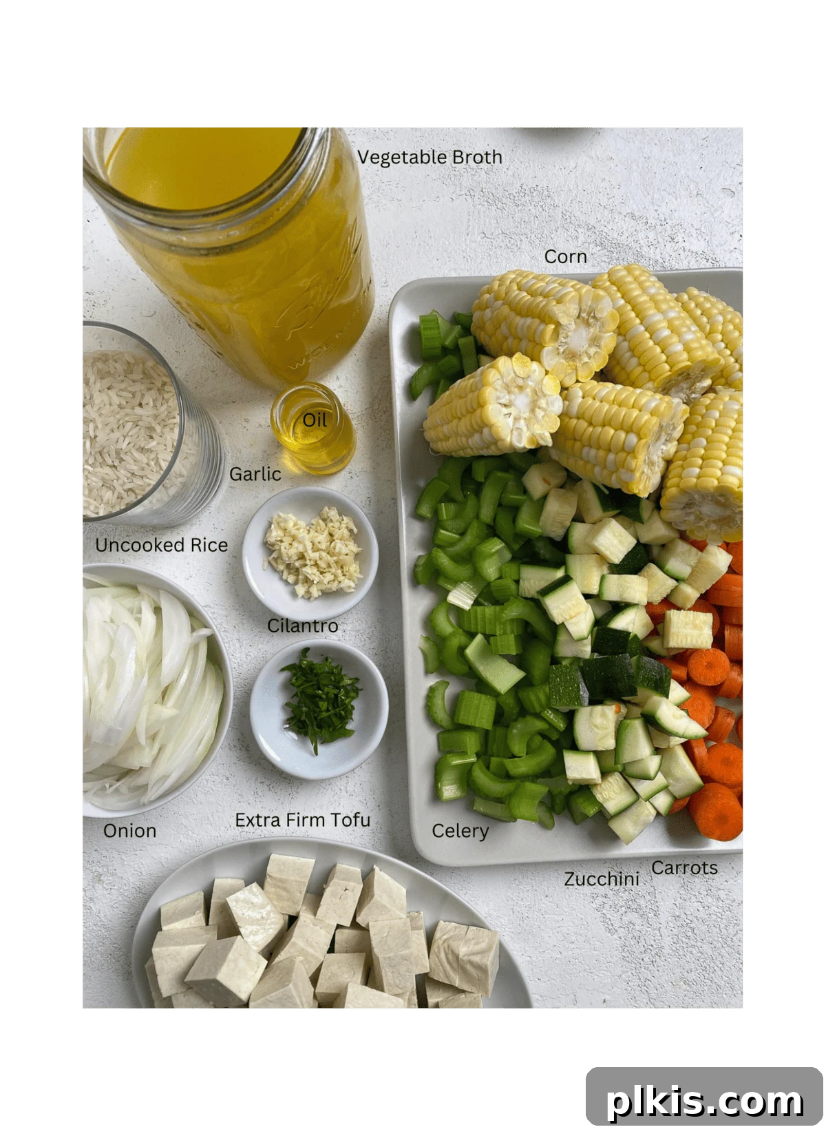 Wholesome Vegan Tofu Broth 3 A selection of fresh ingredients laid out on a white surface, ready for preparing Caldo de Tofu. Included are extra-firm tofu, fresh ears of corn, a zucchini, carrots, celery, yellow onion, garlic cloves, white rice, and a bunch of cilantro.