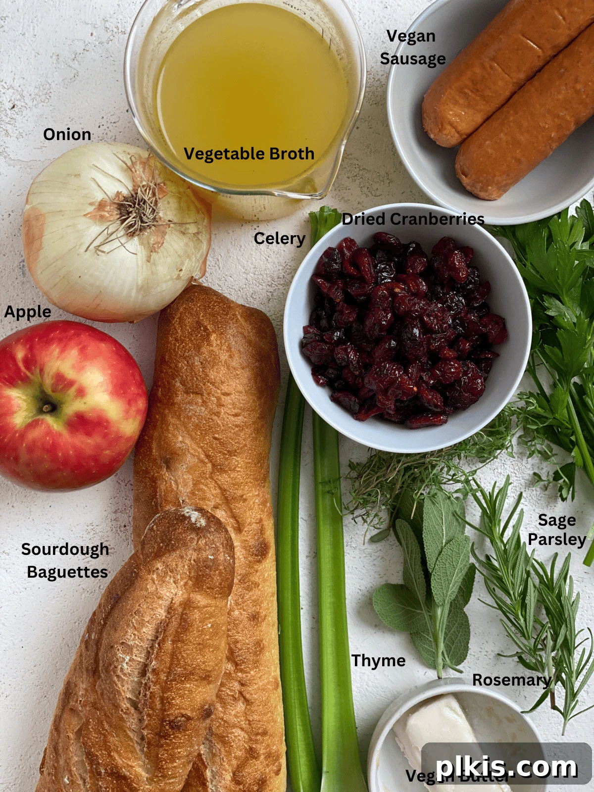 Ultimate Vegan Stuffing 3 Measured out ingredients for The Best Vegan Stuffing on a white surface, including diced sourdough, fresh herbs, cranberries, apples, and vegan sausage.