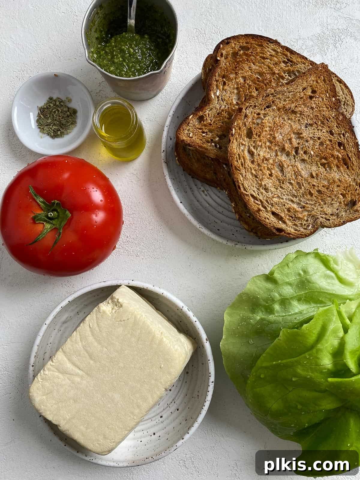 Pesto Tofu Sandwich: Simple Savory Superb 3 All ingredients for a vegan pesto tofu sandwich are neatly measured and arranged on a light surface, including sliced extra-firm tofu, a bottle of olive oil, dried oregano, a jar of pesto, fresh tomatoes, lettuce, and sandwich bread.