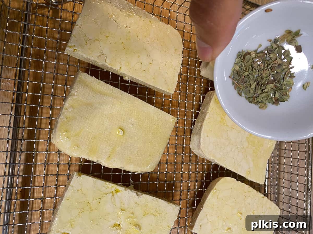 Pesto Tofu Sandwich: Simple Savory Superb 6 Dried oregano being generously sprinkled over olive oil-coated tofu slices on a baking sheet, adding aromatic seasoning.