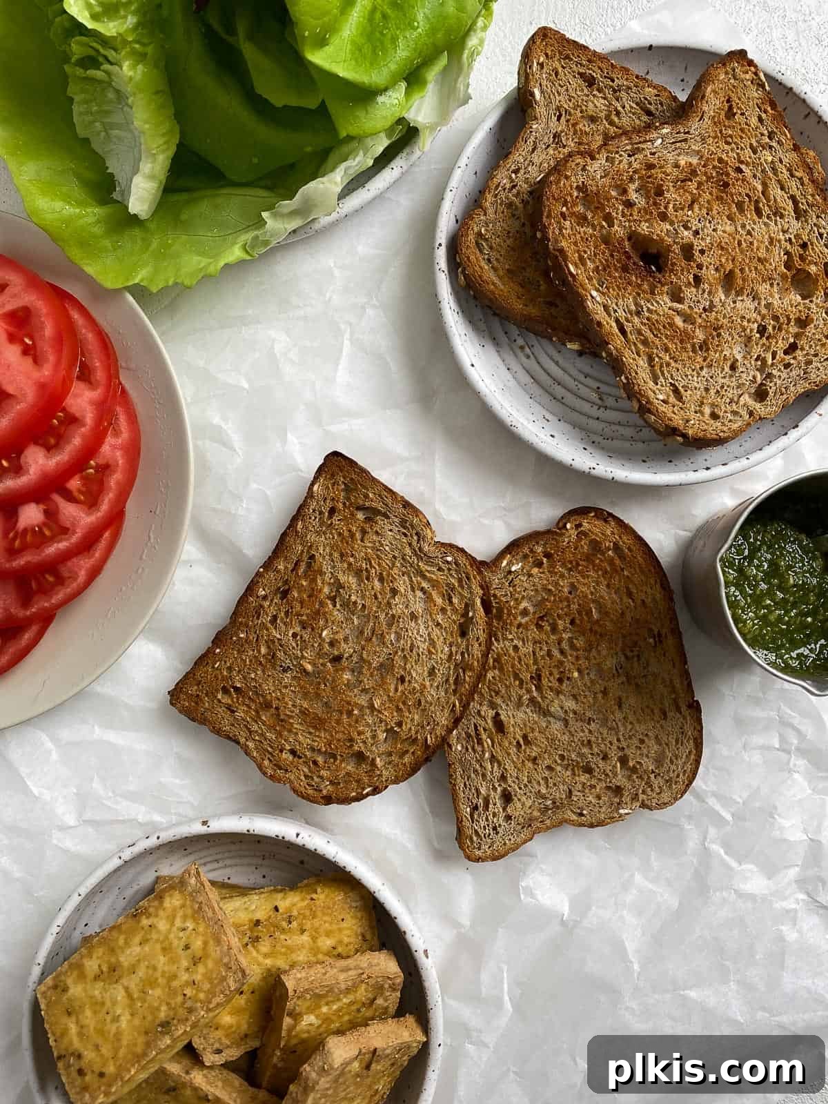 Pesto Tofu Sandwich: Simple Savory Superb 8 Roasted tofu slices displayed alongside fresh ingredients like pesto, lettuce, tomato, and bread, all ready for assembling a delicious vegan pesto tofu sandwich.