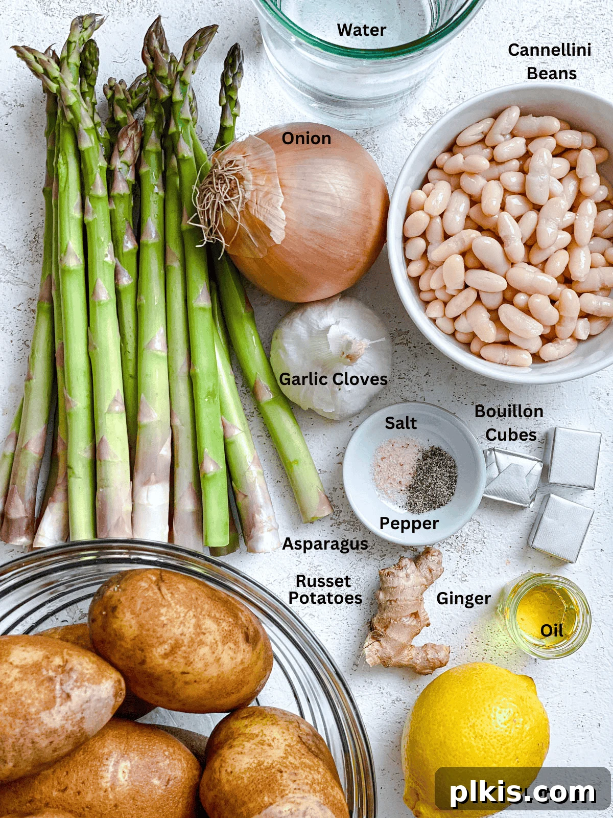 Fresh ingredients for creamy vegan asparagus soup laid out on a white surface, including asparagus, potatoes, white beans, onion, garlic, and lemon.