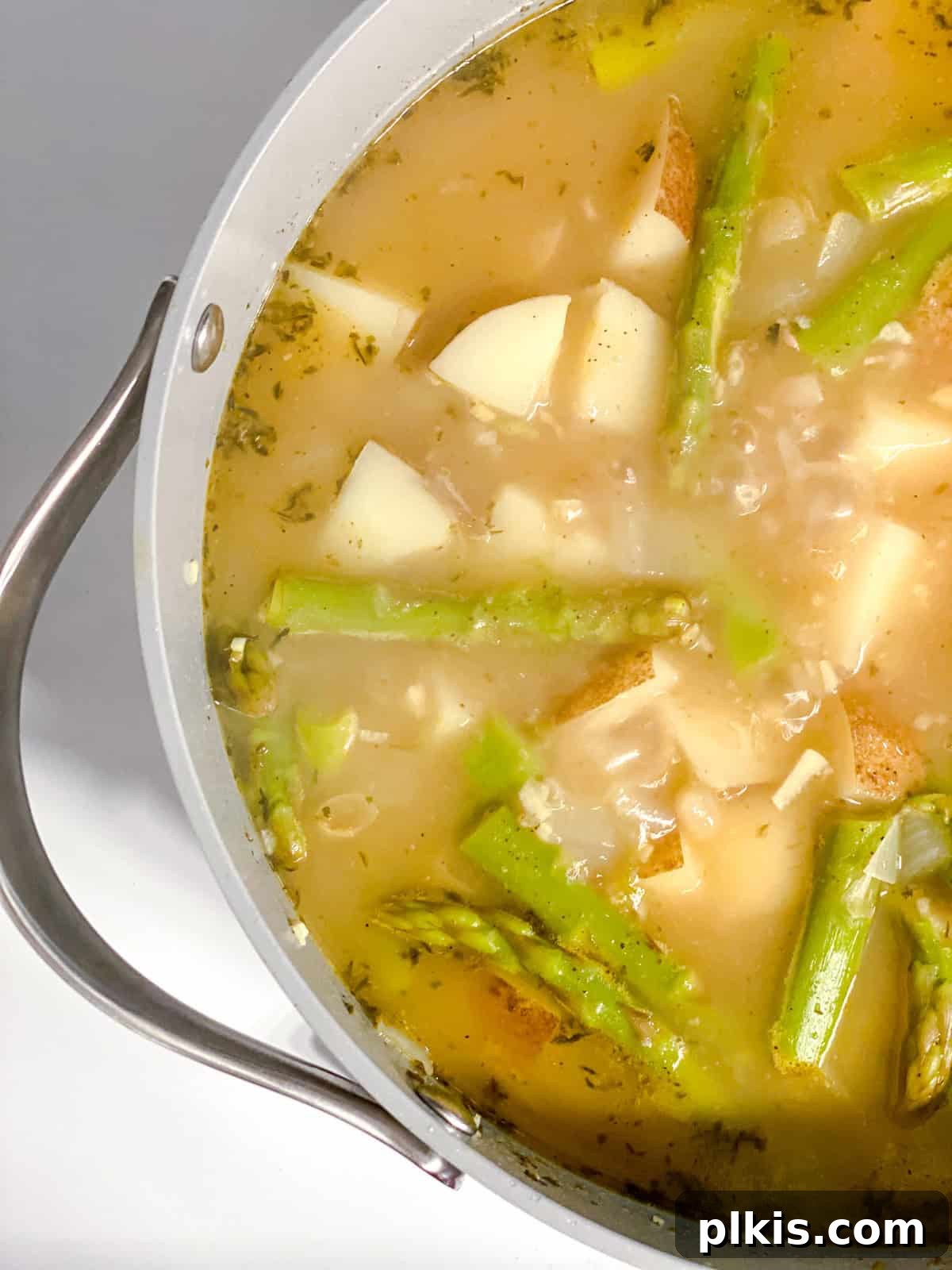 Process shot of potatoes, white beans, and asparagus simmering in a pot of broth, absorbing flavors and softening.