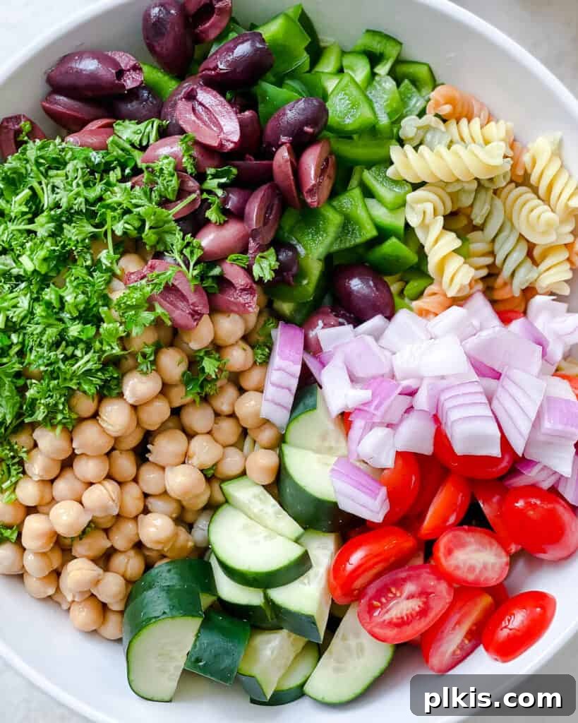 A process shot showing various diced vegetables, chickpeas, and olives being combined in a large mixing bowl.