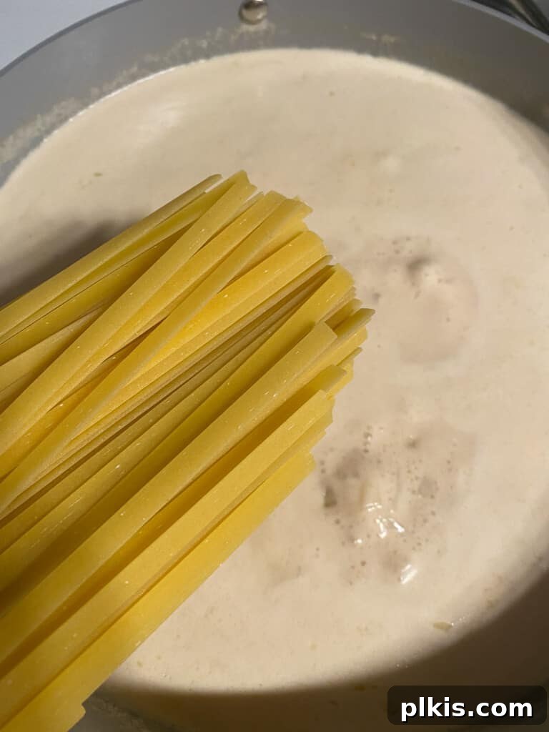 Decadent Vegan Fettuccine Alfredo 7 Process photo showing uncooked fettuccine noodles being added directly into a pot of simmering Alfredo sauce base.