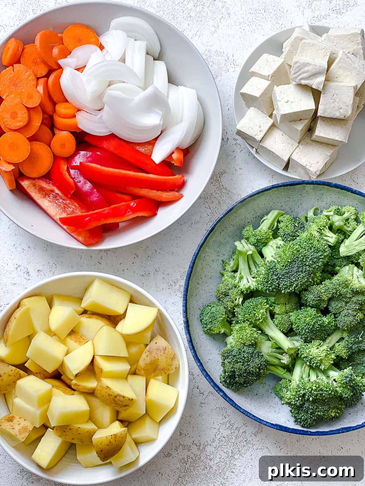 Creamy One-Pot Vegan Yellow Tofu Curry 4 Separately portioned ingredients for the curry: a bowl of chopped potatoes, broccoli florets, sliced red bell peppers, and cubed tofu on a white surface.