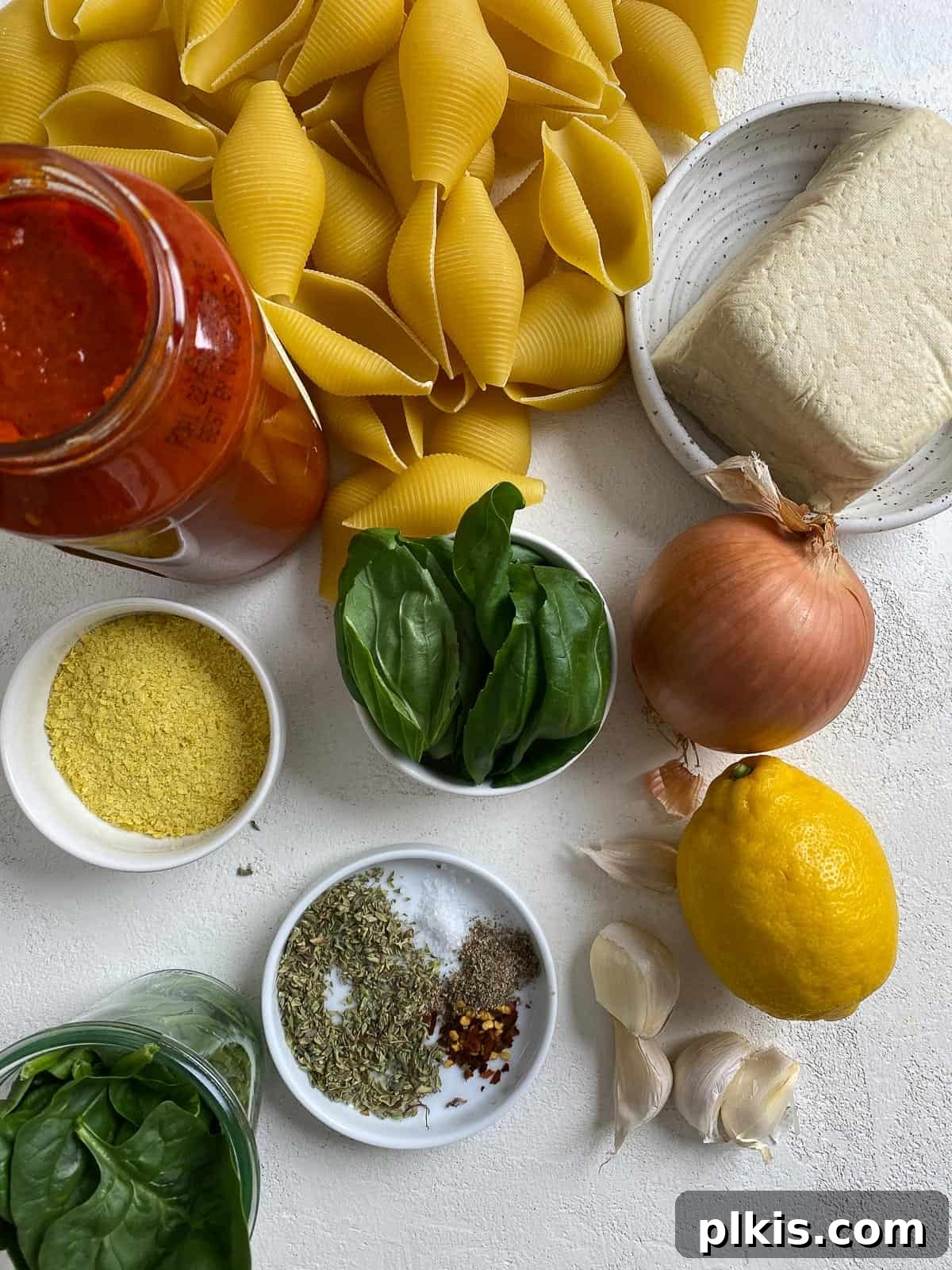 Creamy Vegan Stuffed Shells 4 A selection of fresh and pantry ingredients for Vegan Stuffed Shells, including jumbo shells, tofu, spinach, herbs, and marinara sauce, arranged on a white surface.